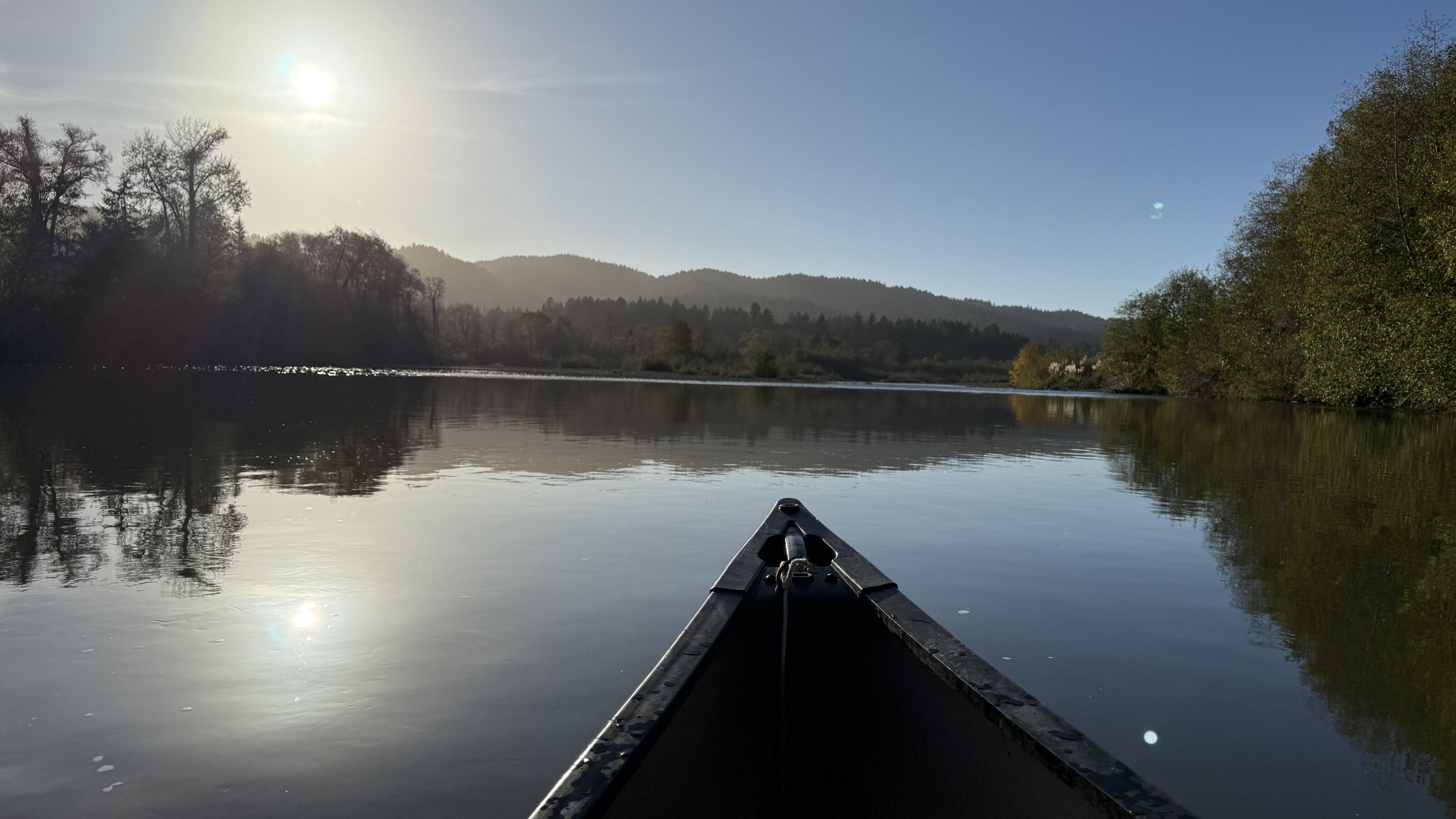 Bow of a canoe gliding across the calm Mad River on a sunlit morning, forested hills on the horizon