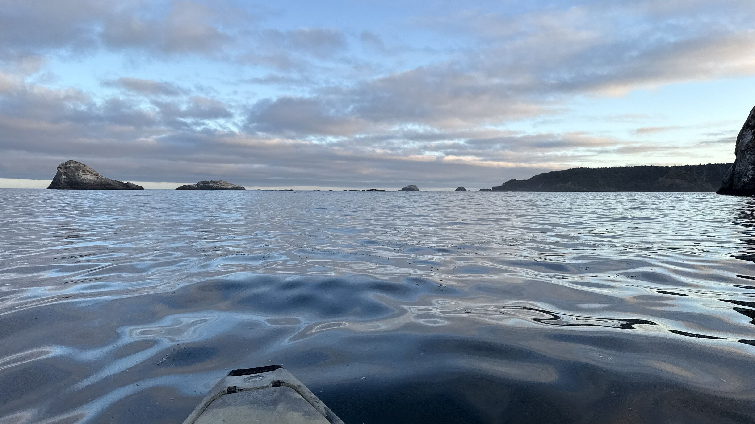 Bow of a kayak on glassy water in Trinidad Bay, sea stacks in the distance under a soft evening sky